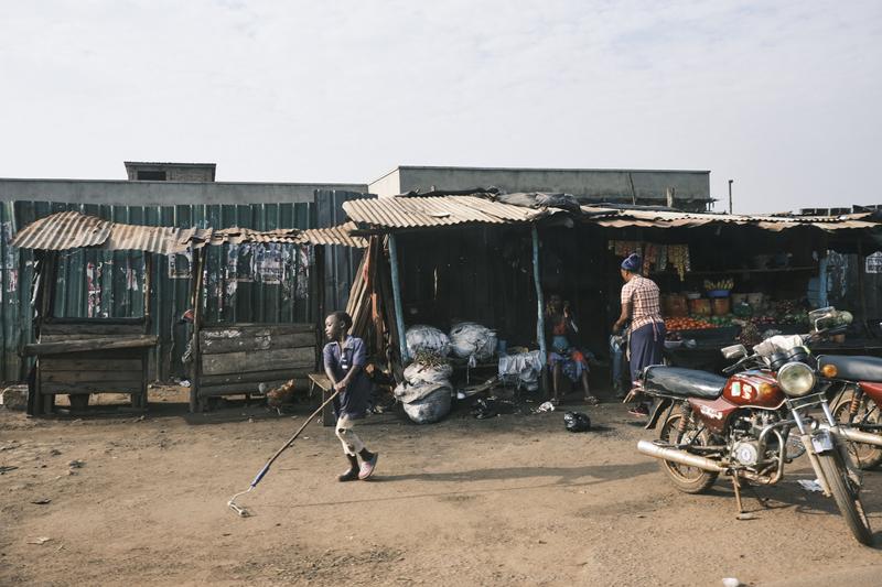 Child playing with a roller by a store front and fruit stand, Uganda